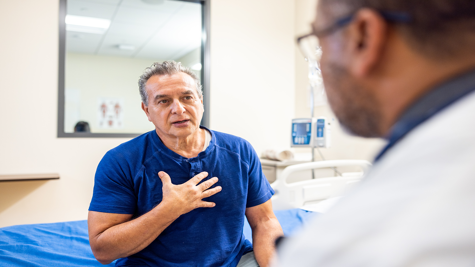 an older man wearing a blue shirt holds his hand over his chest while talking to a doctor about heart failure and health