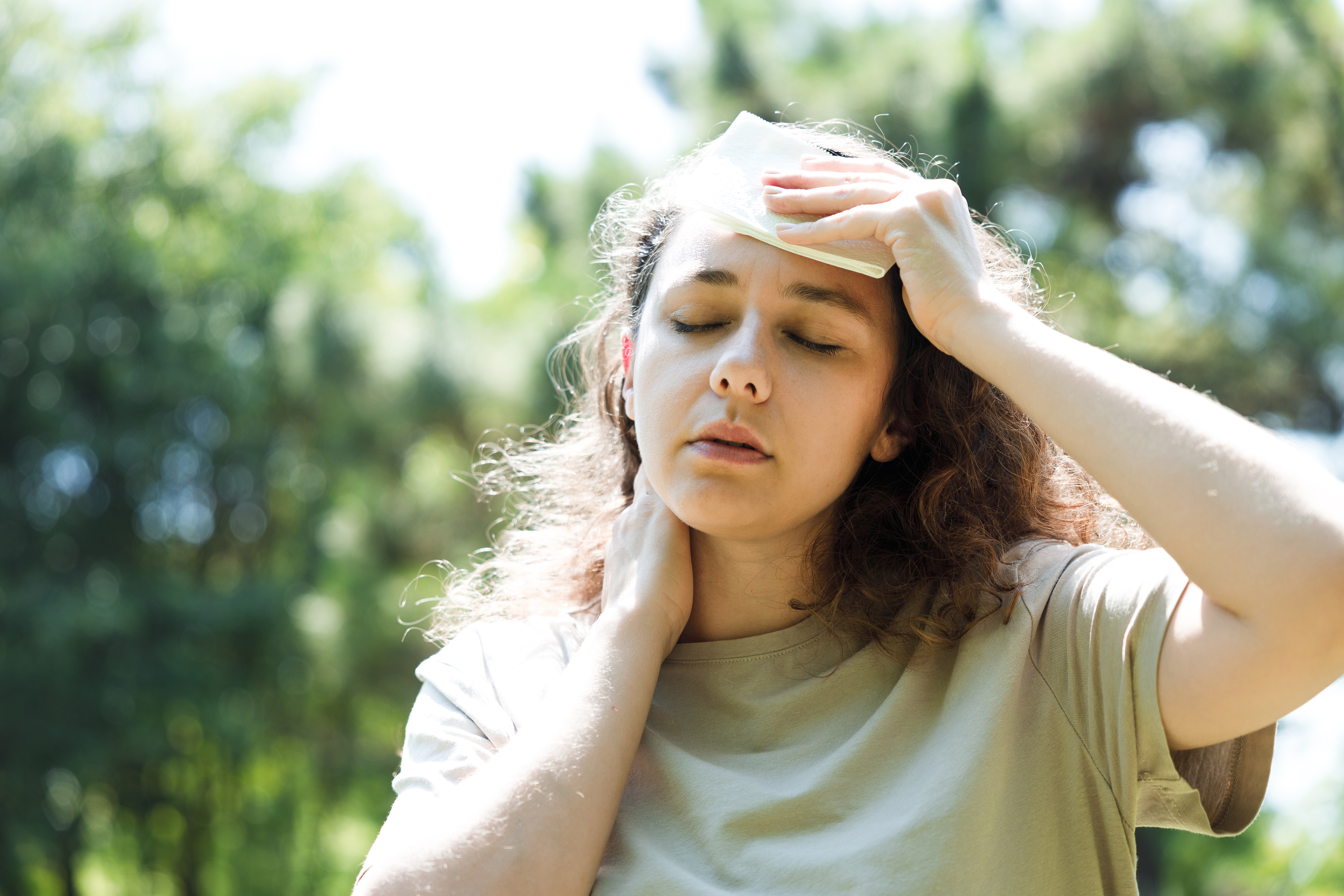 a woman holds a damp cloth to her head while standing outside in the heat, trees seen in the background